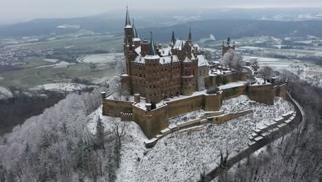 Vuelo-Aéreo-Hacia-Y-Panoramización-Hacia-El-Castillo-Medieval-Hohenzollern-En-Una-Montaña-En-Un-Día-Nevado-Durante-El-Invierno-En-Suabia,-Alemania