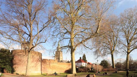 Park-view-time-lapse-of-historic-city-walls-with-stronghold-tower-in-winter-barren-landscape-with-Walburgis-church-tower-of-Zutphen,-The-Netherlands,-in-the-background-against-a-blue-sky-with-clouds