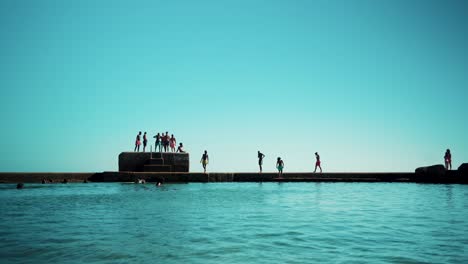 Salt-water-swimming-pool-at-shoreline-with-kids-jumping-from-rock-pier-on-water-at-sunshine-4K
