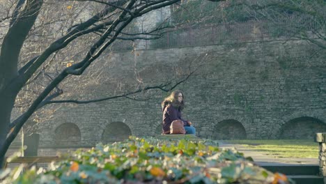 Young-woman-sitting-on-an-old-wall-eats-an-apple-while-looking-at-the-beautiful-and-old-park