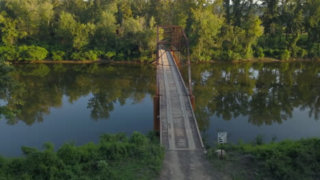 Left-to-right-aerial-slow-truck-shot-of-an-old-iron-bridge-over-a-tree-lined-rural-river-in-warm-late-afternoon-light