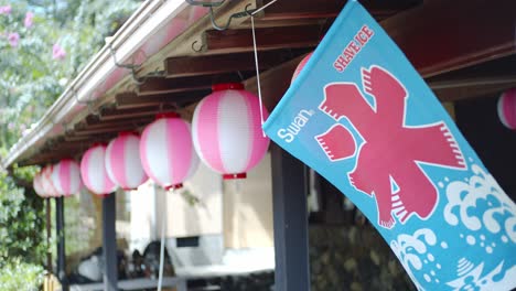 Flag-blowing-in-the-wind-with-lanterns-hanging-on-the-side-of-the-rooftops-of-a-restaurant-in-Kyoto,-Japan-slow-motion-4K