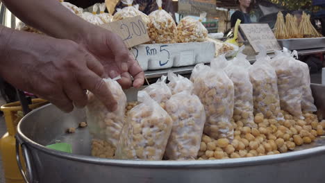 Chickpeas-or-garbanzo-beans-on-display-for-sale-with-man-packing-it-into-clear-small-bag,-price-tag,-popcorn-and-people-in-the-background