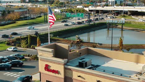 Die-Neigung-Der-Luft-Nach-Oben-Zeigt-Chic-Fil-A-Fastfood,-Amerikanische-Flagge-Und-Stark-Befahrene-Autobahnen