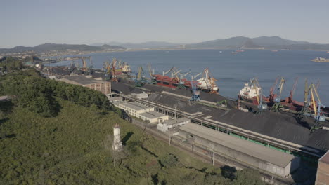 Toma-Aérea-De-Un-Puerto-De-Carbón-Con-Buques-Graneleros-De-Carga-General-Amarrados-Y-Una-Bahía-Con-Una-Colina-Verde,-En-Un-Día-Soleado