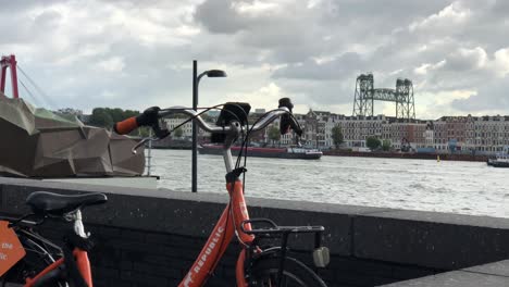 Orange-Donkey-Republic-Fahrrad-Geparkt-Vor-Rotterdamer-Hafen-Mit-Frachtschiff,-Das-Durch-Die-Wasserstraße-Segelt