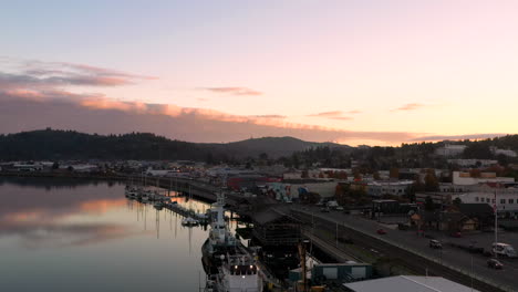 Drone-descends-over-boats-docked-in-Coos-Bay-harbor-during-pink-sunset,-aerial-view-of-city