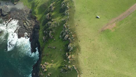 Aerial-top-down,-4x4-drives-close-to-edge-of-ocean-cliffs,-rocky-beach