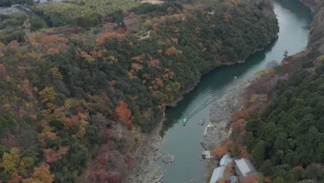 Boat-travels-through-Arashiyama-in-Fall-with-Kyoto-in-the-background,-Aerial-tilt-shot