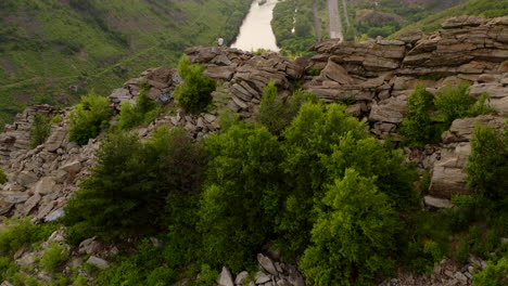 Luftbild-Vor-Einem-Jungen-Mann,-Der-Auf-Einer-Felsigen-Klippe-In-Pennsylvania-Sitzt-Und-Den-Grünen-Wald-Und-Den-Fluss-Während-Eines-Sommersonnenuntergangs-Zeigt