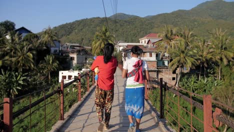 Following-women-carrying-child-walking-over-high-suspension-bridge-crossing-in-tropical,-sunny,-Muang-Khua-,-northern-Laos