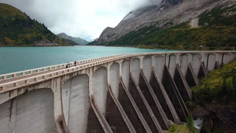 Lake-Fedaia-Dam-in-the-Dolomite-mountain-range-with-people-admiring-the-view-from-the-road-on-the-structure,-Aerial-Drone-flyover-view