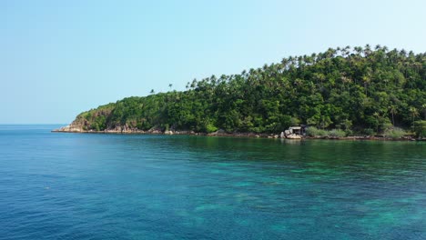 Tranquil-lagoon-with-coral-reefs-growing-under-crystal-water-near-rocky-coastline-of-tropical-island-with-trees-forest-hills-in-Thailand