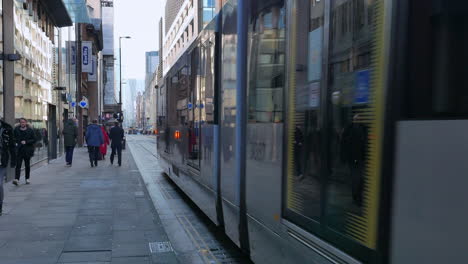 Gente-Caminando-Por-Una-Calle-En-Manchester-Con-Un-Tranvía-Bajando