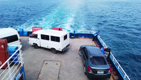 Vista-Superior-De-La-Cubierta-De-Un-Gran-Barco-Que-Transporta-Camiones-Con-Gente-Mirando-El-Océano-Mientras-El-Barco-Está-En-Marcha-Sobre-Un-Océano-Azul