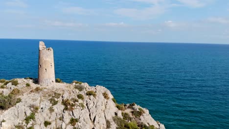 Drone-flies-over-a-coastal-watchtower,-centennial-tower-on-the-blue-sea-cliff-and-rocks-in-a-beautiful-aerial-view