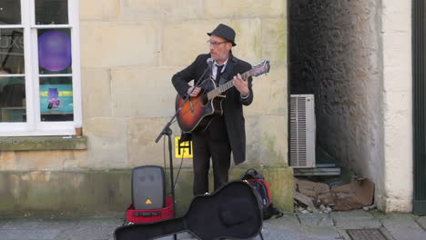 Old-Slim-Man-Wearing-Black-Suit-Strumming-Guitar-And-Singing-Outside-in-Truro,-Cornwall---Wide-Shot