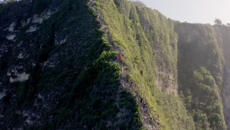 Kelingking-Beach-T-Rex-peninsula-with-people-descending-cliff-staircase,-Aerial-approach-shot