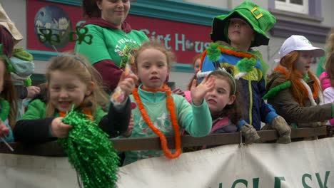 Grupo-De-Niños-Parados-En-Vagones-De-Camiones-Vestidos-De-Verde,-Usando-Símbolos-Irlandeses,-Tréboles-Y-Sombreros-Verdes,-Saludando-A-La-Cámara-Y-Sonriendo