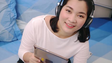 Schöne-Asiatische-Frau-Schaut-Kamera-Und-Lächelt,-Entspannt-Sich-Und-Hört-Musik-Per-Tablet-Mit-Kopfhörern-Auf-Dem-Bett-Zu-Hause-Lifestyle--Und-Vergnügungskonzept