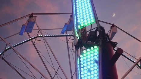 Satellite-ride-at-carnival-spinning-clockwise-with-moon-and-ferris-wheel-in-background-at-night