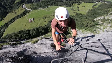 A-young-white-man-is-happy-and-smiling-as-he-climbs-up-the-ladder-made-out-of-small-metal-handles-on-the-steep-cliff-of-a-mountain-in-Furenalp,-Ferrata-in-Switzerland
