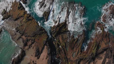Top-down-zoom-towards-the-ground-in-Porto-das-Salemas-with-the-waves-of-the-turquoise-water-crashing-on-the-rocky-shore