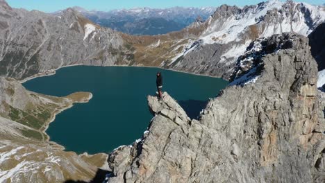 Circling-drone-shot-of-a-young-man-standing-in-a-black-jacket-and-shorts-on-the-top-of-a-cliff,-overlooking-the-heart-shaped-lake-and-the-mountain-landscape-by-himself-in-the-sun