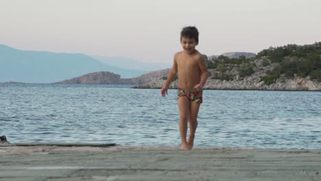 Caucasian-toddler-boy,-running-happily-on-a-dick-next-to-the-sea,-wearing-a-swimsuit,-slow-motion-120fps