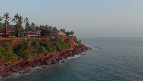 Aerial-view-of-Varkala-Beach,-Kerala,-India-showing-resorts-and-cafes-on-dense-green-cliffs-along-the-seashore-with-low-tides-hitting-them