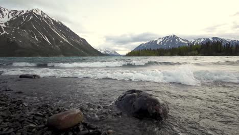 Low-angle-view-of-Yukon-Kathleen-lake-waves-rolling-onto-rocky-shore-with-scenic-snow-capped-mountains-in-background,-Canada,-wide-angle-static