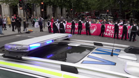 Ein-Polizeiauto-Mit-Blaulicht-Ist-Auf-Der-Straße-Vor-Den-Umweltprotestierenden-Der-Politischen-Partei-Geparkt,-Die-Eine-Straßenkreuzung-Auf-Dem-Trafalgar-Square-Mit-Einem-Großen-Hellrosa-Banner-Blockieren