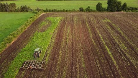 Tractor-Tilling-The-Wide-Field-In-Monroe-County-Michigan-Preparation-For-A-New-Farm-Garden-Background-With-Green-Grass,-Trees,-And-Variety-Of-Plants-On-A-Sunny-Day---Wide-Shot