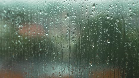 Rain-drops-sliding-slow-on-window-glass-in-rainy-day,-de-focused-city-in-background,medium-closeup-shot