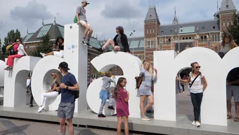 Slow-motion-pan-to-sky-past-groups-of-people-sitting-and-standing-with-I-Amsterdam-sign,-Amsterdam,-Netherlands
