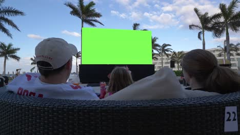 Family-of-3-watching-large-outdoor-green-screen-at-a-tropical-resort