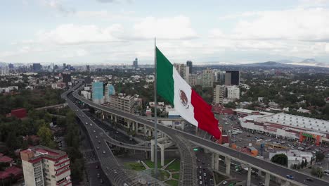 Circle-flight-by-large-majestic-patriotic-red,-white-and-green-Mexican-flag-waving-in-wind-above-Periferico-sur-street-in-downtown-Mexico-city-center,-San-Jeronimo,-on-cloudy-sky-day,-aerial