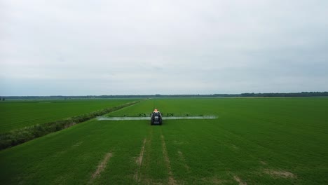 Aerial-view-of-tractor-spraying-pesticide-and-fertilizer-on-the-green-field-in-overcast-summer-day,-wide-angle-ascending-drone-shot-mowing-forward