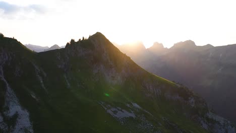 Aerial-shot-flying-sideways-to-the-right,-revealing-the-hot,-warm,-glowing-sun-during-a-sunrise-or-sunset,-peeking-through-behind-the-peaks-of-the-mountains-in-Switzerland