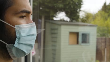 Male-looking-out-of-a-window-longingly-wearing-a-facemask-in-quarantine-close-up