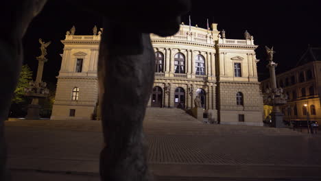 The-maginificent-concert-hall-of-Rudolfinum-with-bright-facade-in-Prague,-Czechia-on-an-empty-square-of-paved-stones-at-night,-during-a-Covid-19-lockdown,-shot-from-behind-a-statue-and-its-legs