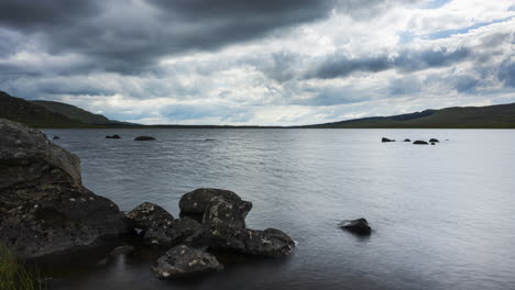 Time-lapse-of-lake-with-grass-and-large-rocks-in-the-foreground-on-a-dark-cloudy-summer-day-in-rural-landscape-of-Ireland