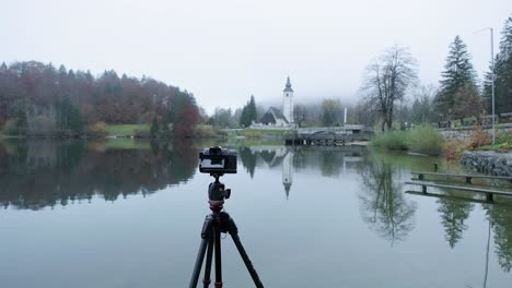 A-camera-on-a-tripod-at-Lake-Bohinj-photographing-a-picturesque-church-with-a-stunning-reflection-in-foggy-conditions-Photography-at-Lake-Bohinj-Church-with-low-clouds