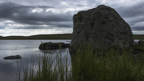 Time-lapse-of-lake-with-grass-and-large-rocks-in-the-foreground-on-a-dark-cloudy-summer-day-in-rural-landscape-of-Ireland