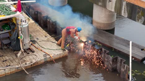 A-man-cutting-a-vertical-line-in-sheet-pile-using-a-blow-torch,-on-a-small-float,-over-water