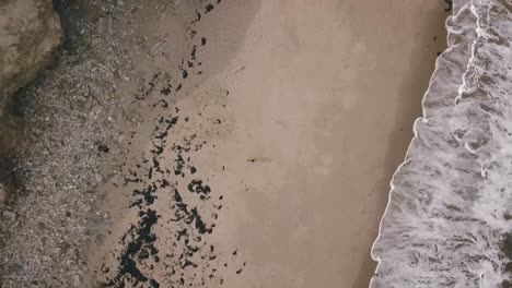 Aerial-birds-eye-view-drone-footage-of-a-man-on-an-empty-beach-with-big-ocean-sea-waves-crashing-onto-the-shoreline