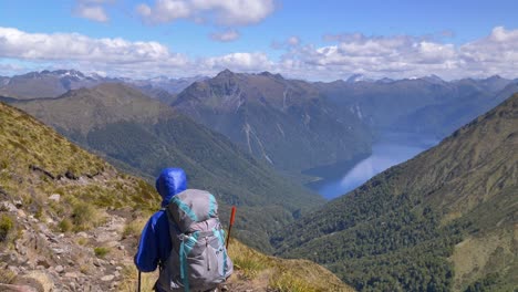 Static,-hiker-walks-along-exposed-alpine-path,-distant-lake-and-mountain-landscape,-Kepler-Track-New-Zealand
