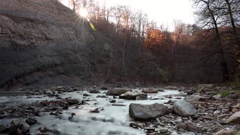 Time-lapse-view-of-a-rocky-river-in-middle-of-leafless,-foliage-forest,-sunny,-fall-evening