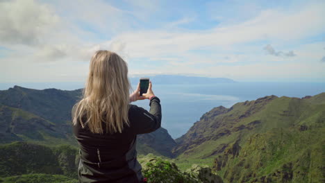 Back-View-Of-A-Caucasian-Woman-Taking-Photo-Of-Majestic-Mountain-With-A-Smartphone-On-A-Sunny-Day,-medium-shot