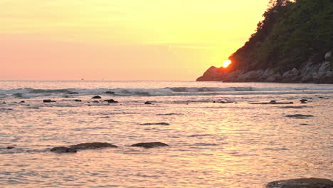 Rocky-sea-shore-during-sunset-and-sun-disk-hides-behind-the-island-mountain-in-Thailand